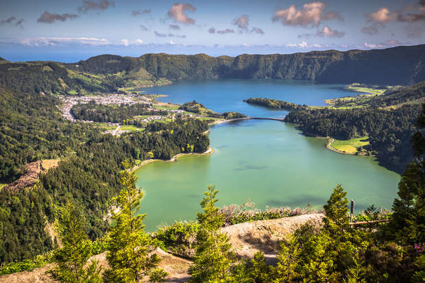 Lakes are surrounded by lush green hills and scattered houses under a partly cloudy sky, with a diverse landscape combining forested areas and open fields adding depth to the view.